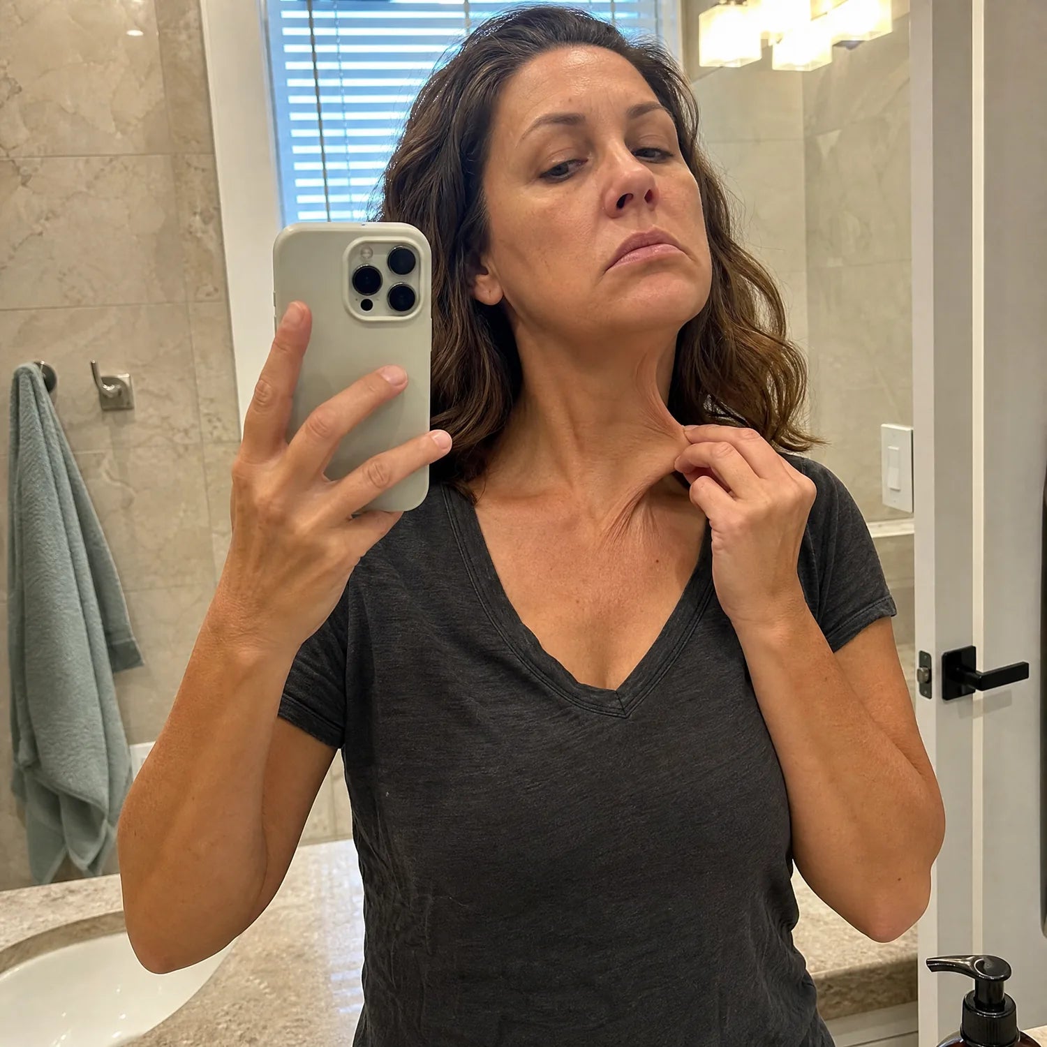 Woman in her late 40s in a bathroom mirror selfie tilting her head up · fingers gently pulling the loose skin under her jawline outward to show visible neck-laxity and turkey-neck-lines · concerned pensive expression · charcoal V-neck t-shirt · sharp focus throughout
